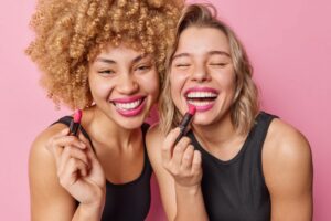 Two women laughing while applying bright pink lipstick